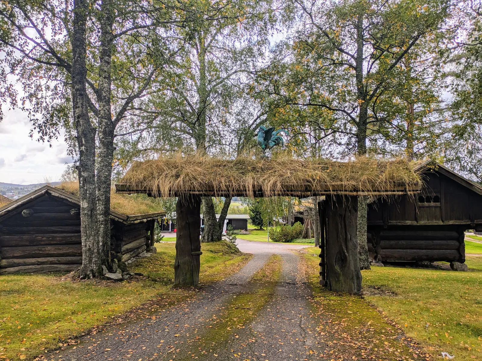 The empty turf-roofed timber gateway over the gravel road into the farm, between birches with their first yellow leaves and log storehouses on either side.